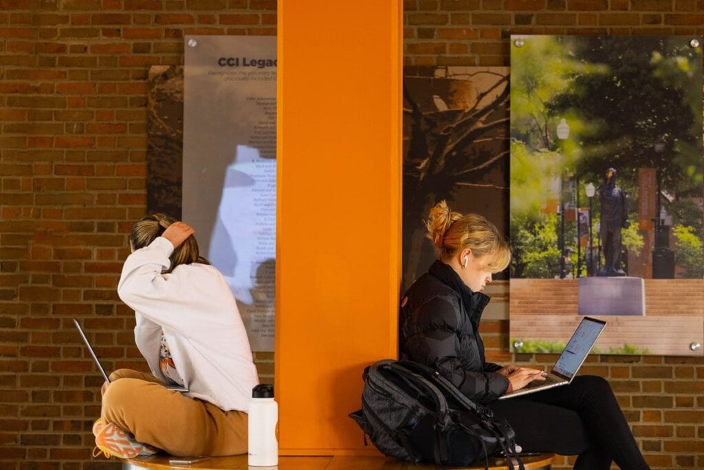 Two students lean their backs against an orange pillar while working on their laptops.